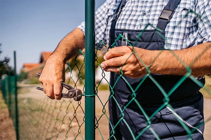 A worker re-tying chain-link fence fabric to a post during a DIY fence repair